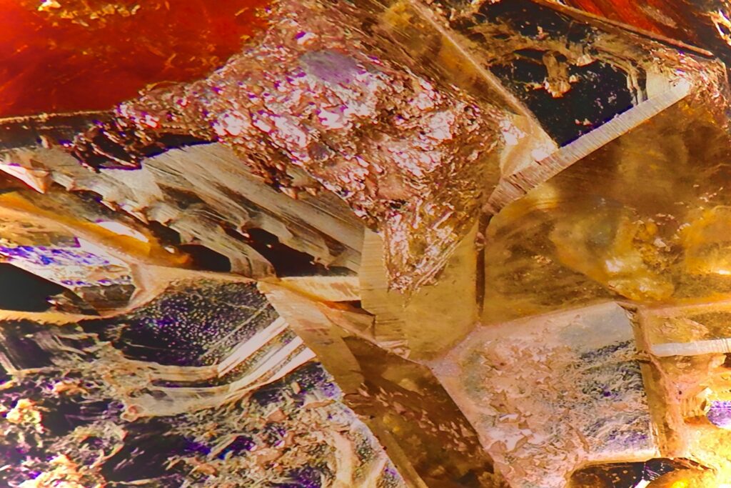 A macro photograph of tightly packed, fractured and faceted mineral pieces. Translucent golden-yellow and golden-brown crystals are jumbled with a large section of rough red mineral and pockets of deep purple and metallic-flecked stone, creating a vibrant, detailed abstract mosaic. The light catches the multiple sharp and rough surfaces.