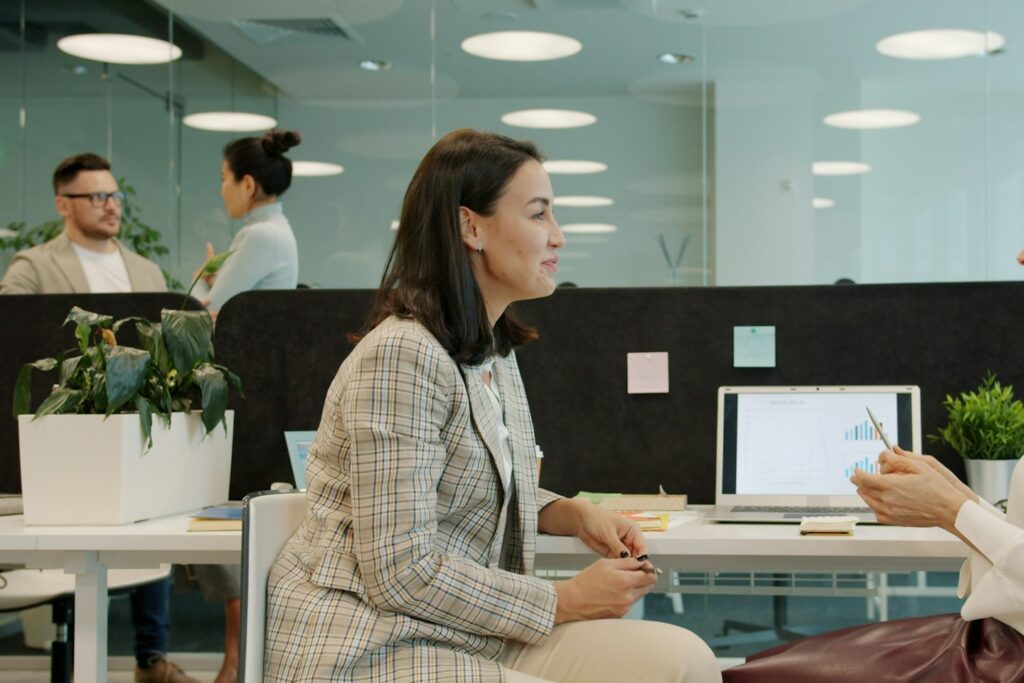 Cheerful women coworkers talking discussing work looking at laptop screen sitting at desk in open space office room. People and communication concept.